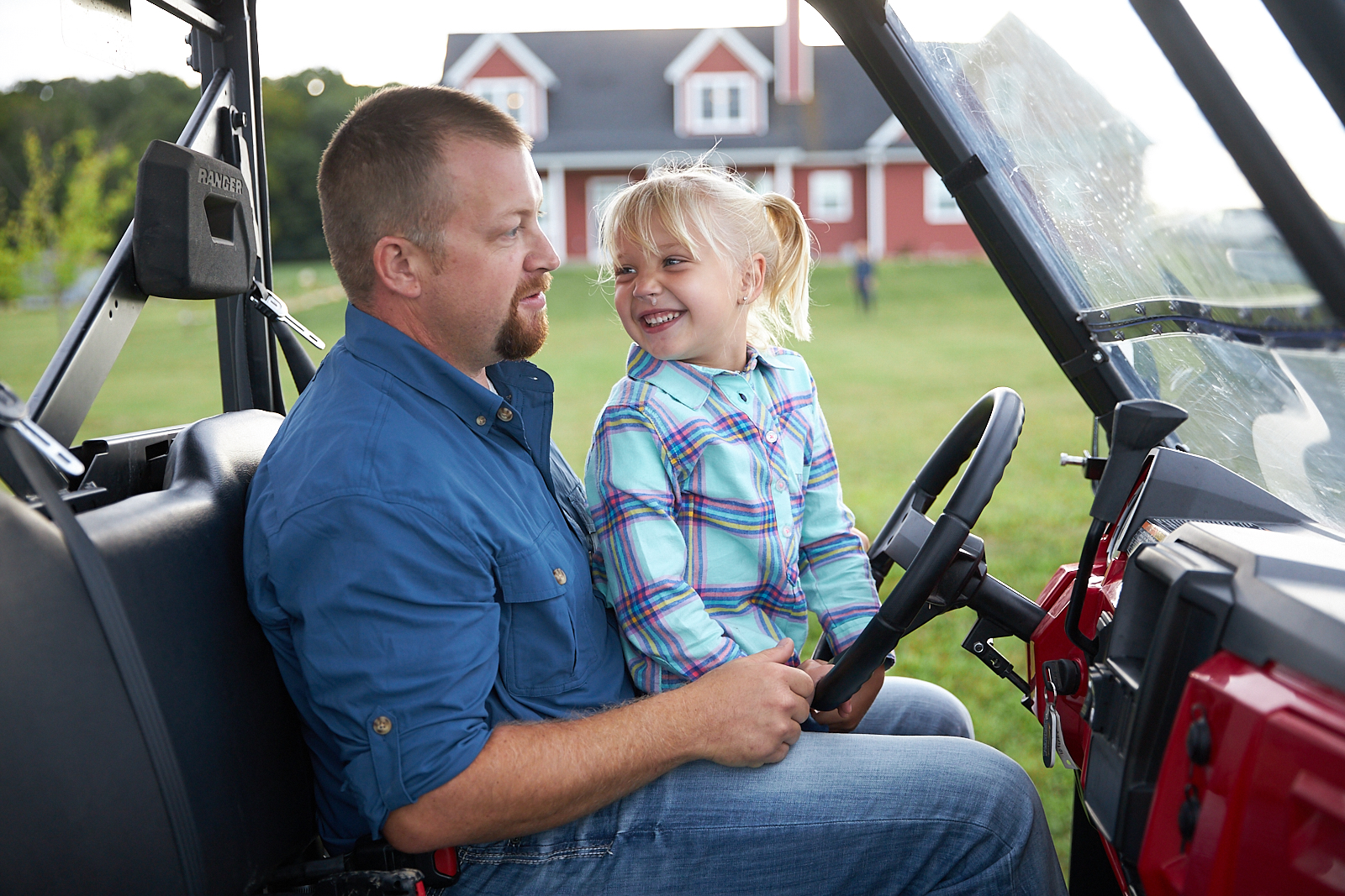 Father and daughter smile in a utility vehicle near a red farmhouse.