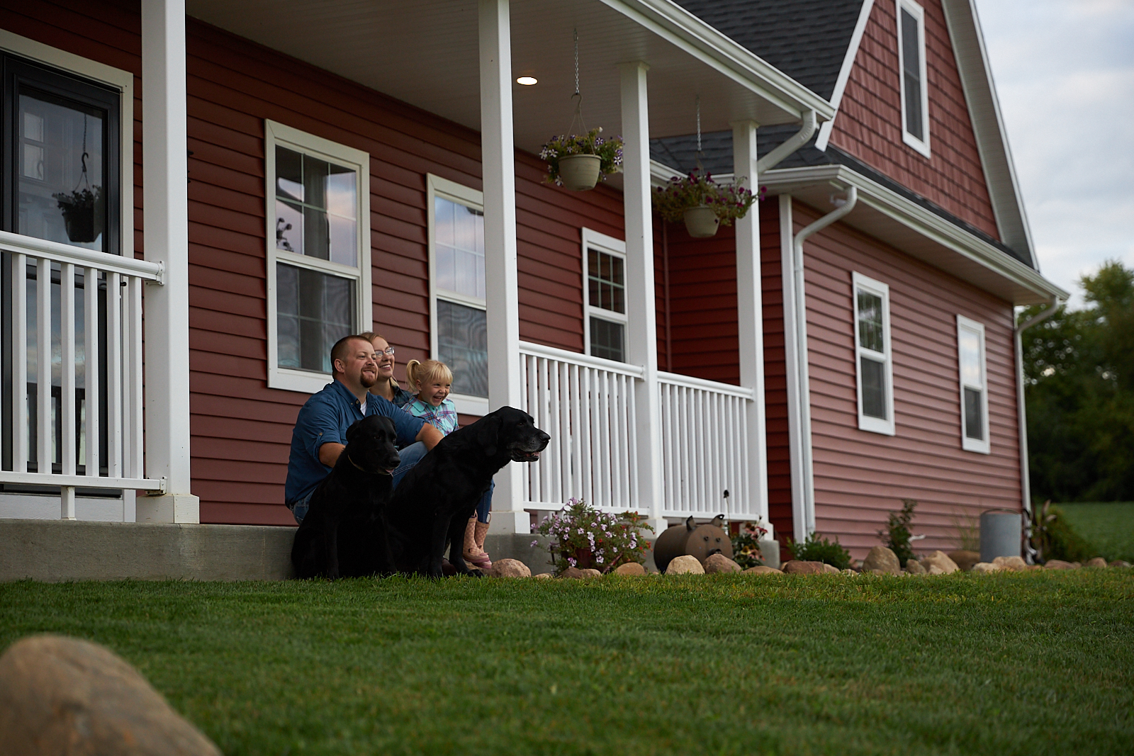 Family with two black dogs sitting on porch of red farmhouse.