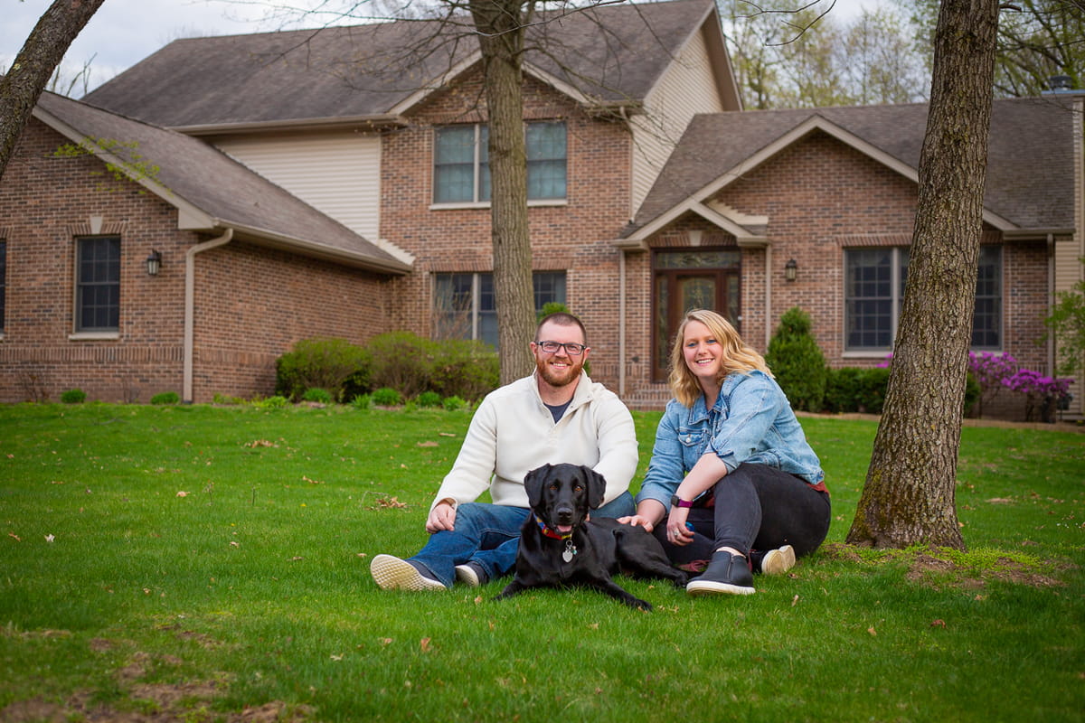 Couple sitting on the grass with their dog in front of a brick home surrounded by trees.