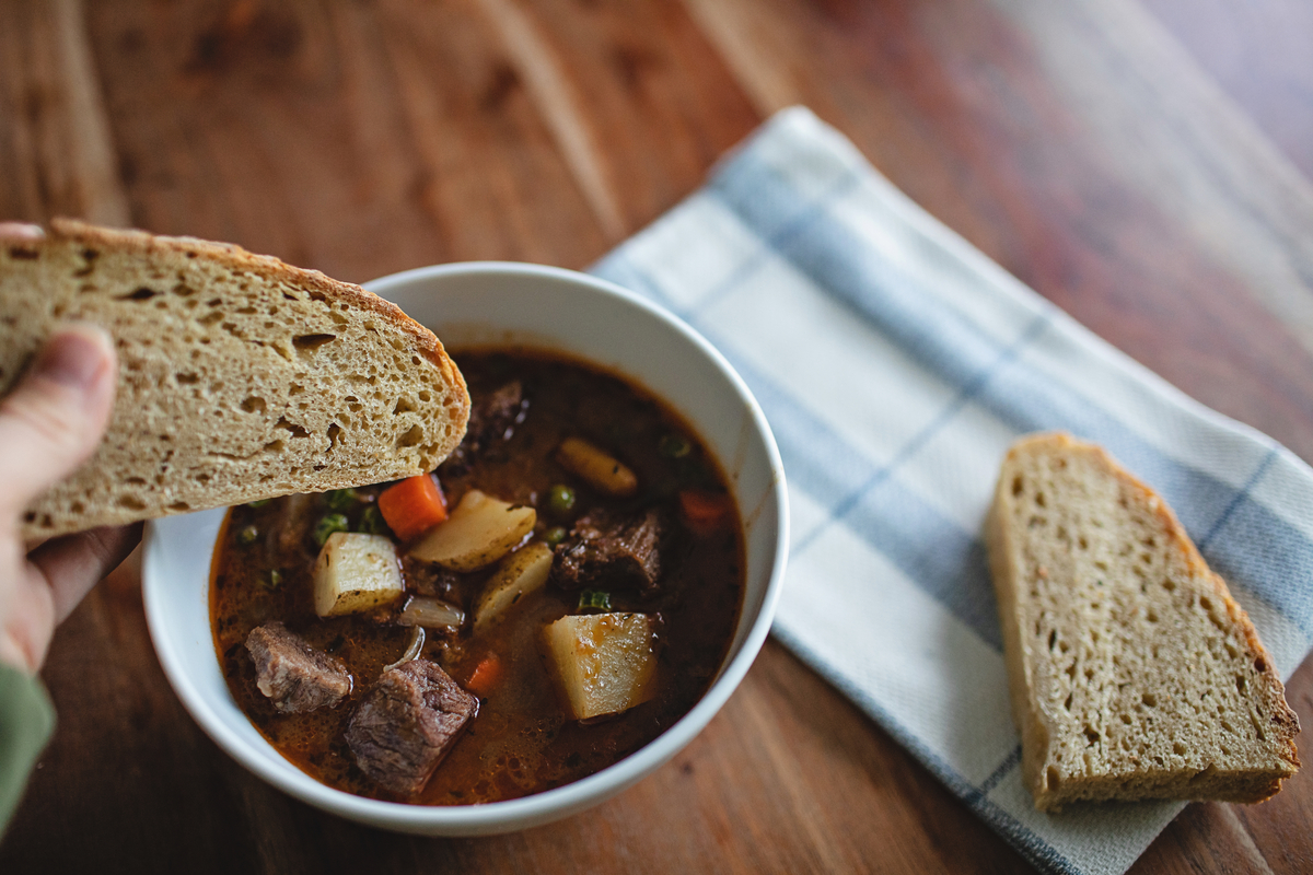 Bowl of beef and vegetable stew with a hand holding a slice of rustic bread above it on a wooden table. 