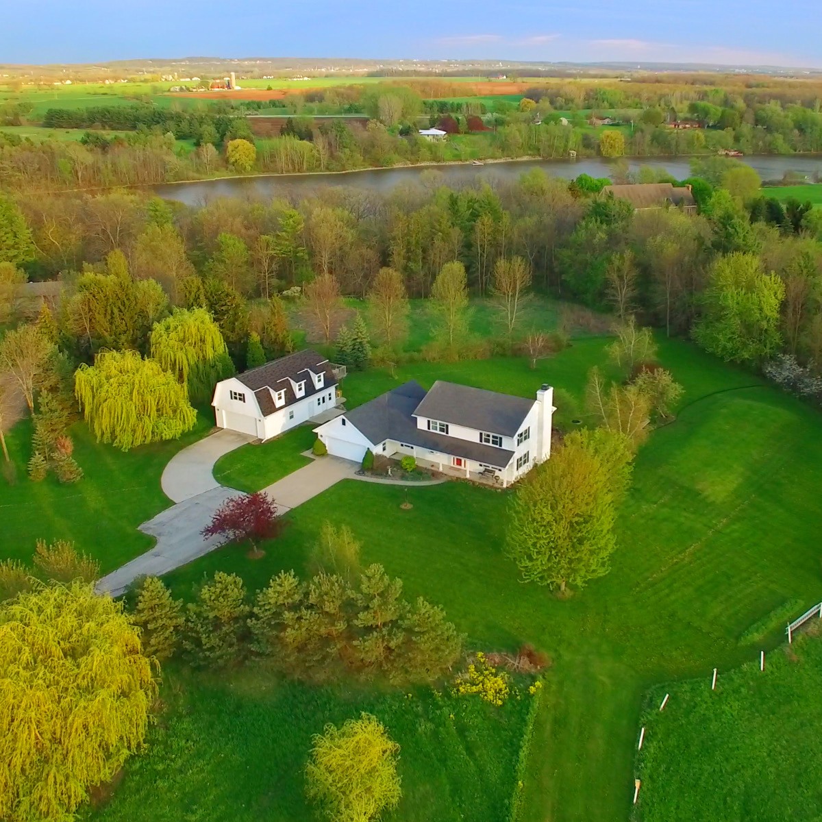 An aerial view of a white rural house, surrounded by lush green trees, tall grass and a blue sky.