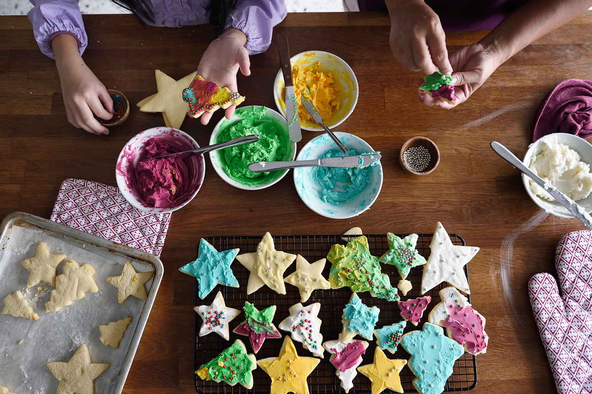 : Colorful holiday cookies cooling on a rack surrounded by bowls of frosting and sprinkles.