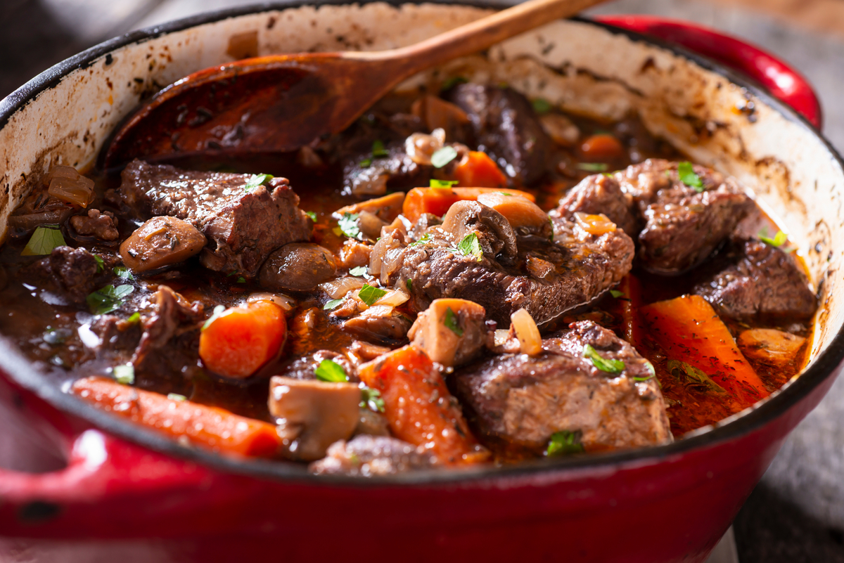 Close-up of a red Dutch oven filled with beef stew, showing tender chunks of meat, carrots, mushrooms, and herbs simmering together.
