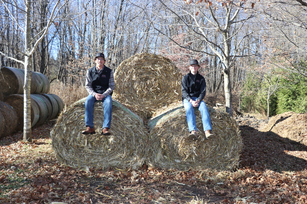 Two young men wearing vests and hats sit on stacked hay bales in a wooded rural setting on a sunny day.