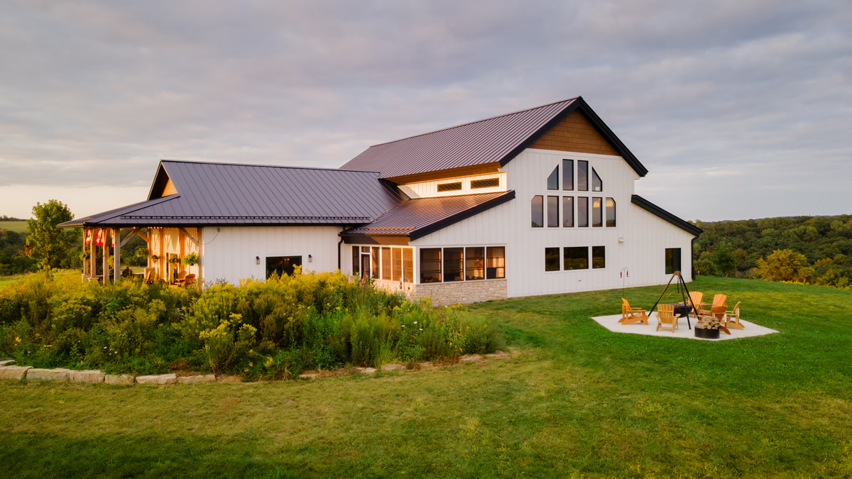 Modern white barndominium surrounded by greenery, lit by warm sunset light.