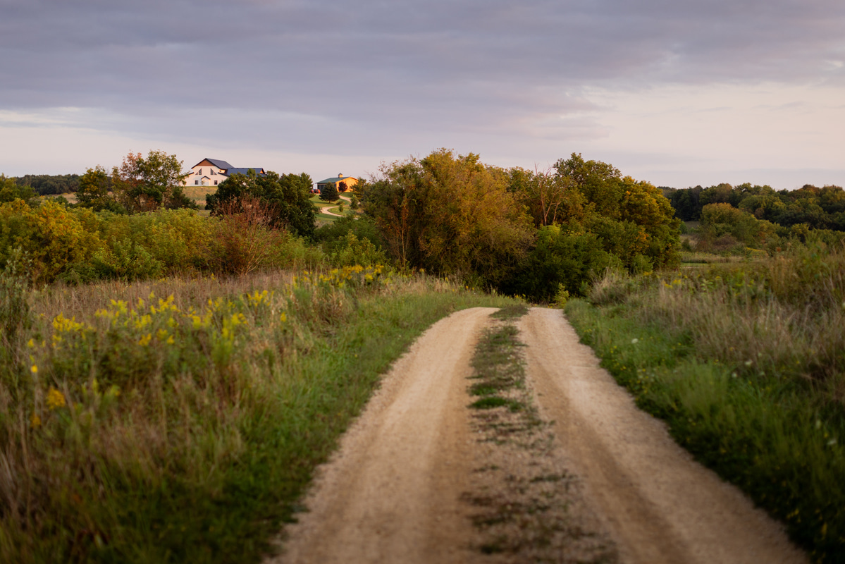 Gravel driveway leading to a countryside barndominium surrounded by trees at sunset.