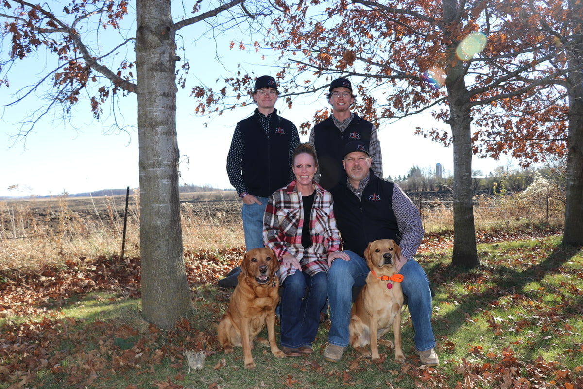 Family poses outdoors under autumn trees with two dogs on a rural farm property.
