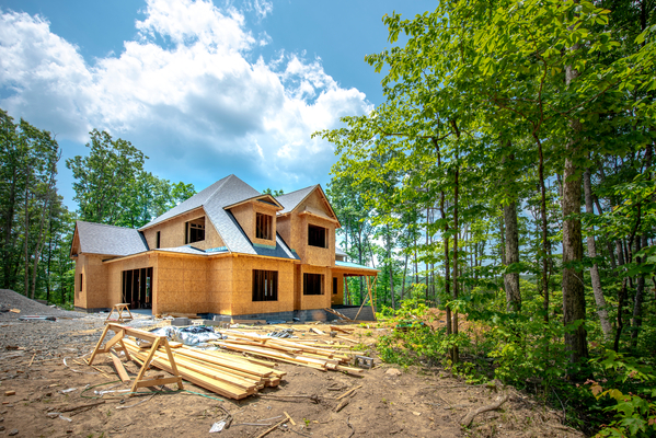 A large two-story house under construction with OSB sheathing stands in a wooded clearing under a bright, cloudy sky.
