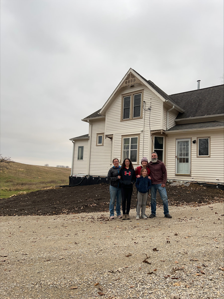 Family standing together outside a white farmhouse on a gravel driveway under cloudy skies.