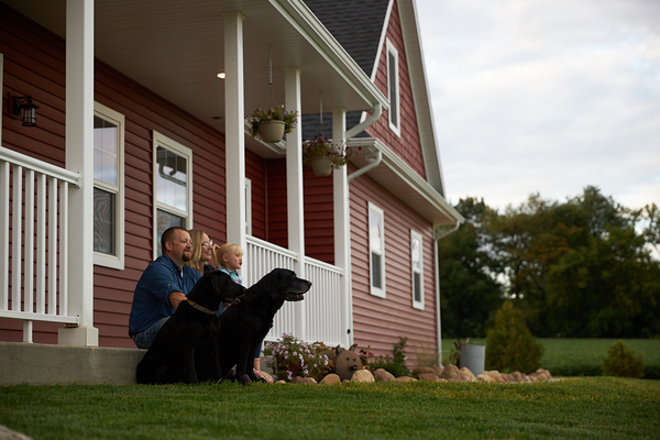 Family with two black dogs sitting on porch of red farmhouse.