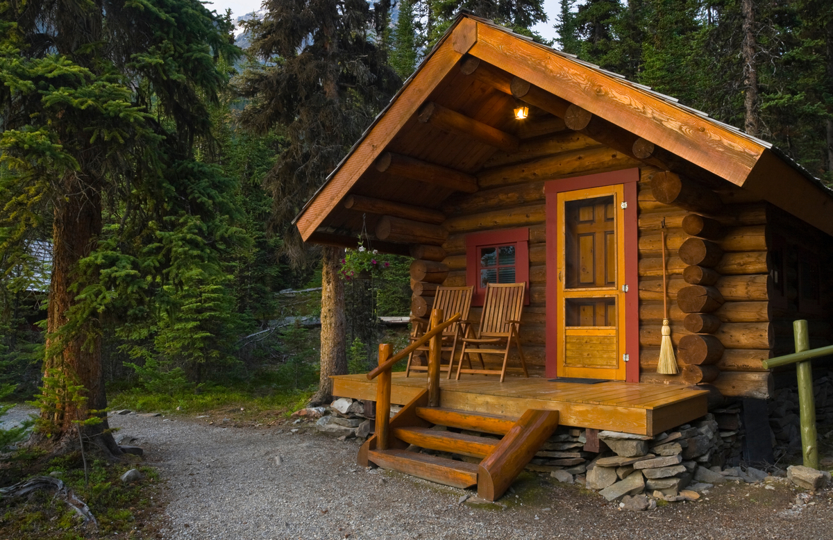 Cozy log cabin with front porch and chairs surrounded by dense forest scenery.