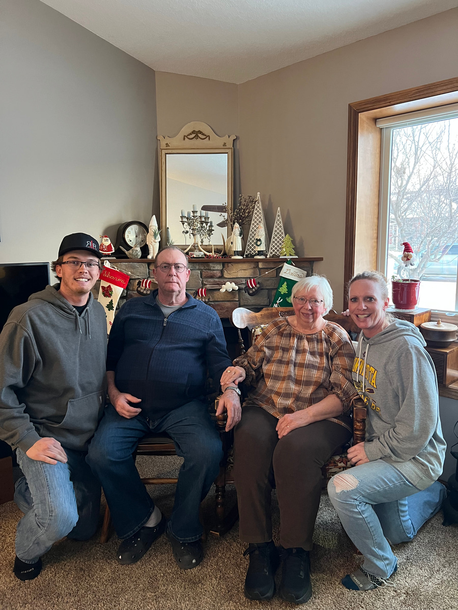 Four adults seated together in a cozy living room, smiling in front of a fireplace decorated with holiday stockings and winter-themed décor.
