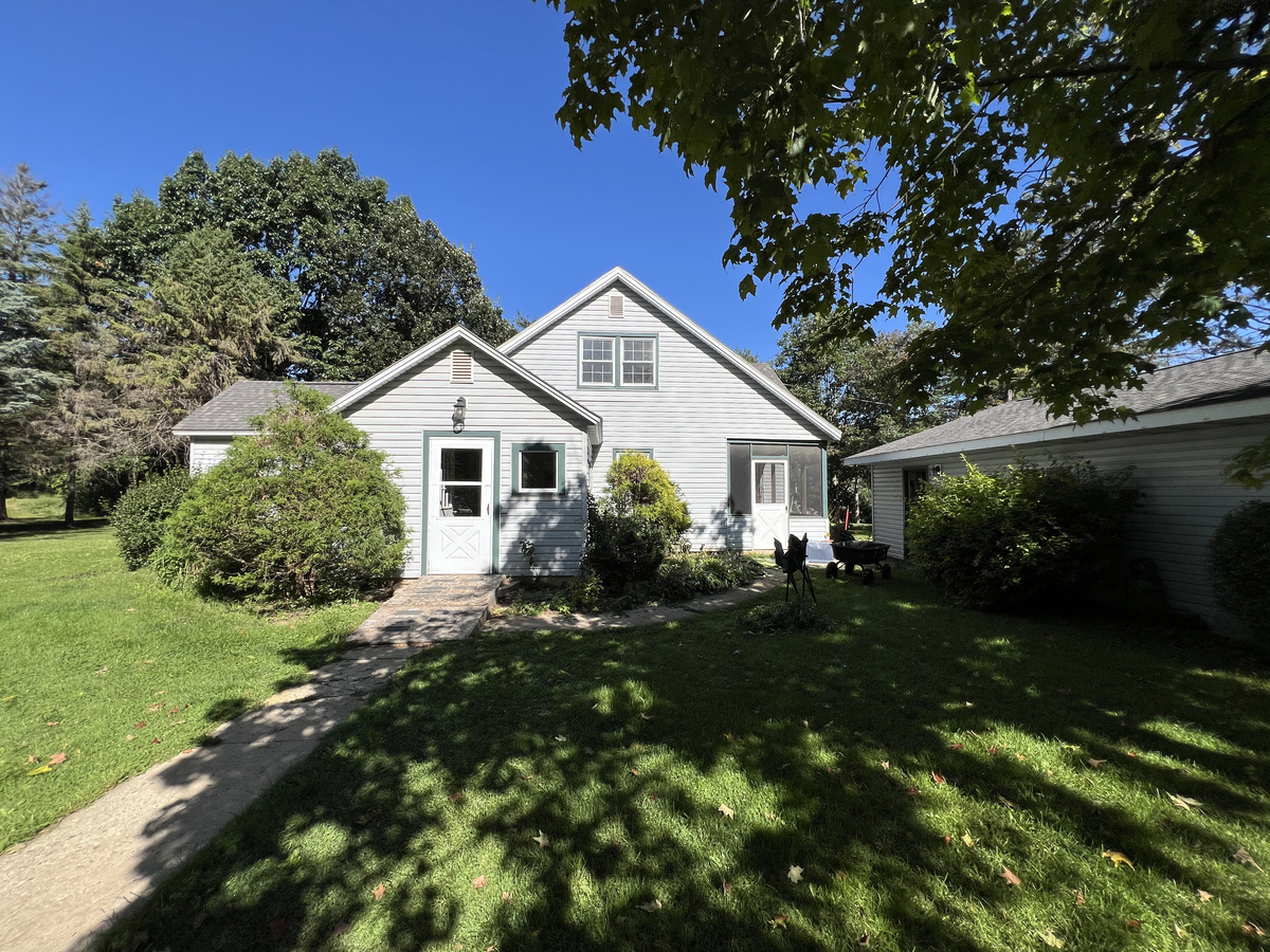 Gray farmhouse with white trim sits among mature trees with a detached garage and green lawn on a sunny day.