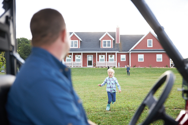 Father and daughter smile in a utility vehicle near a red farmhouse.