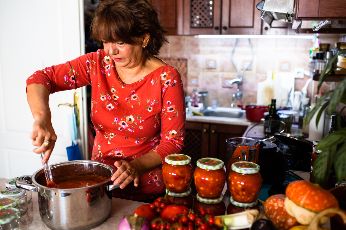 Woman stirring tomato sauce in kitchen with jars of homemade preserves on the counter.