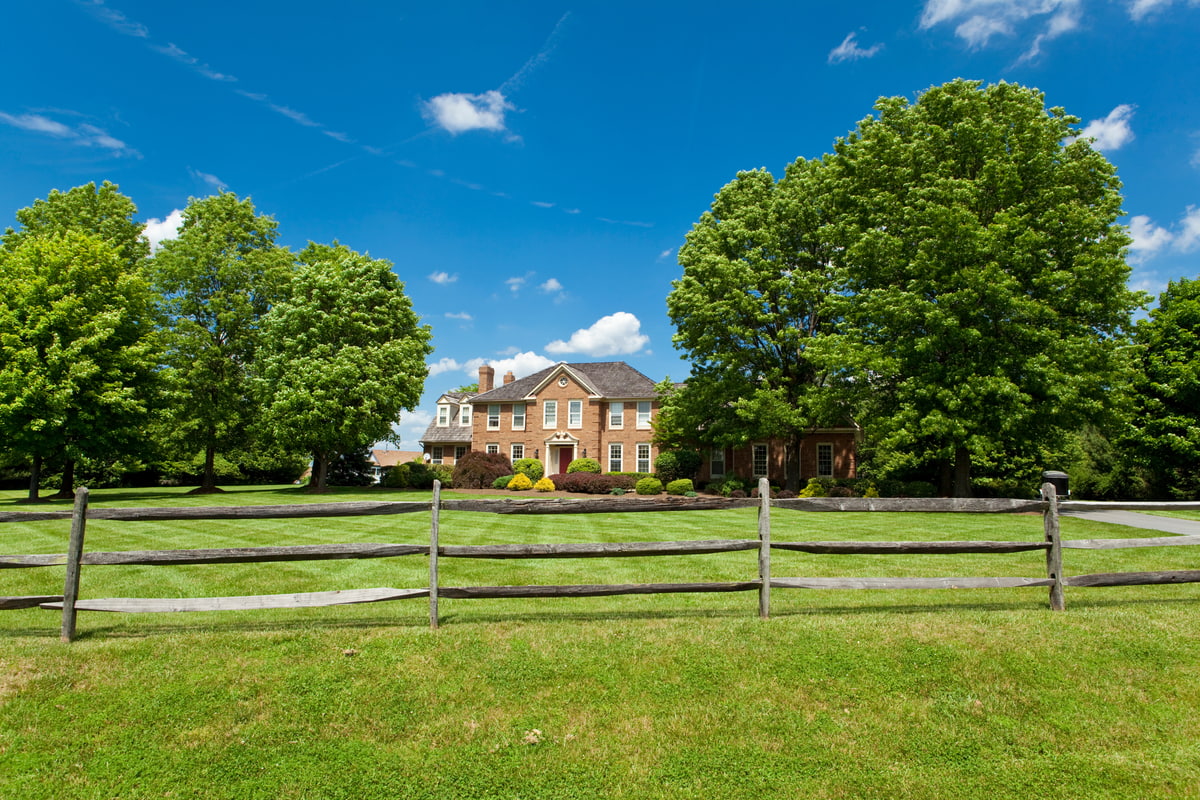 A large brick home with white trim and a red door sits behind a wooden fence, surrounded by lush trees and a manicured lawn under a bright blue sky.