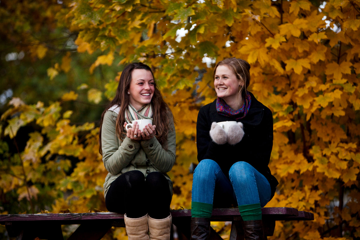 Two women sitting outside in autumn, smiling and holding mugs of apple cider.