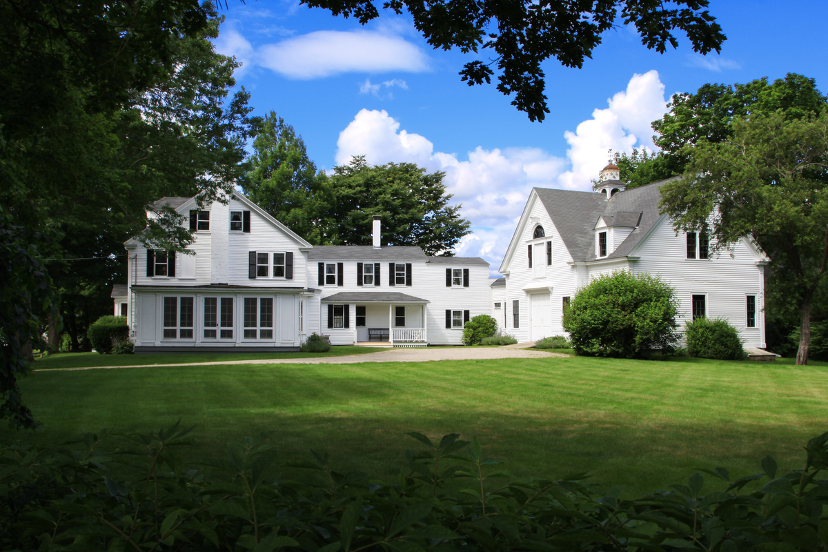 Large white farmhouse with manicured green lawn and trees under blue sky.