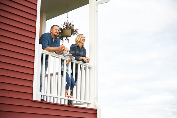 Parents talk on farmhouse porch overlooking fields while child rests in arms.