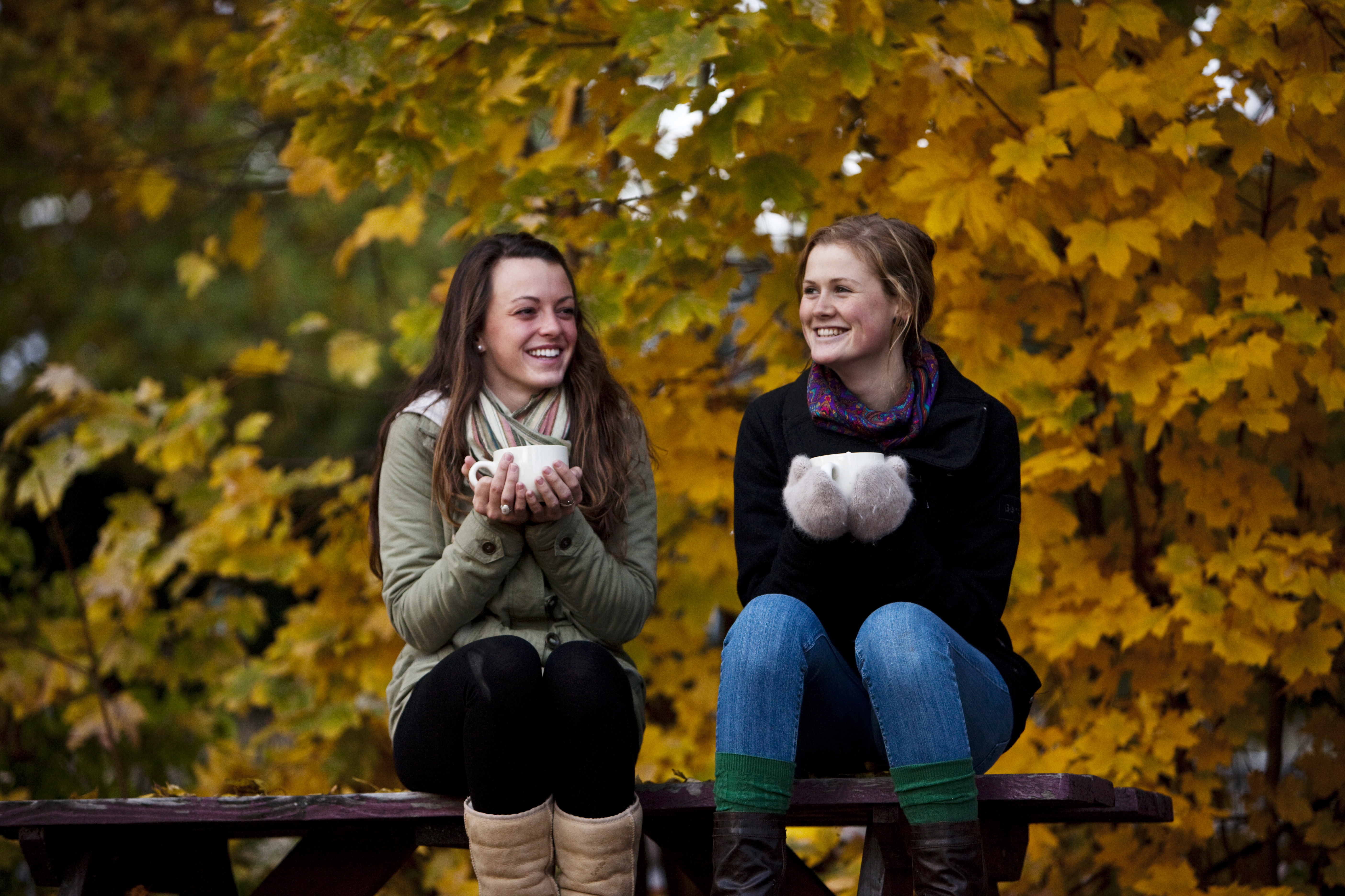 Two women sitting outside in autumn, smiling and holding mugs of apple cider.