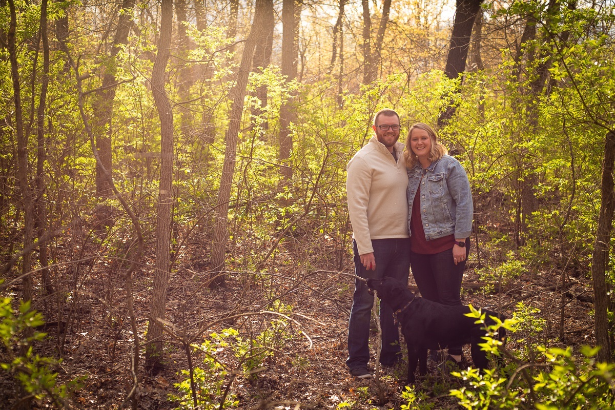 a couple stands in sunlit foliage with their black dog
