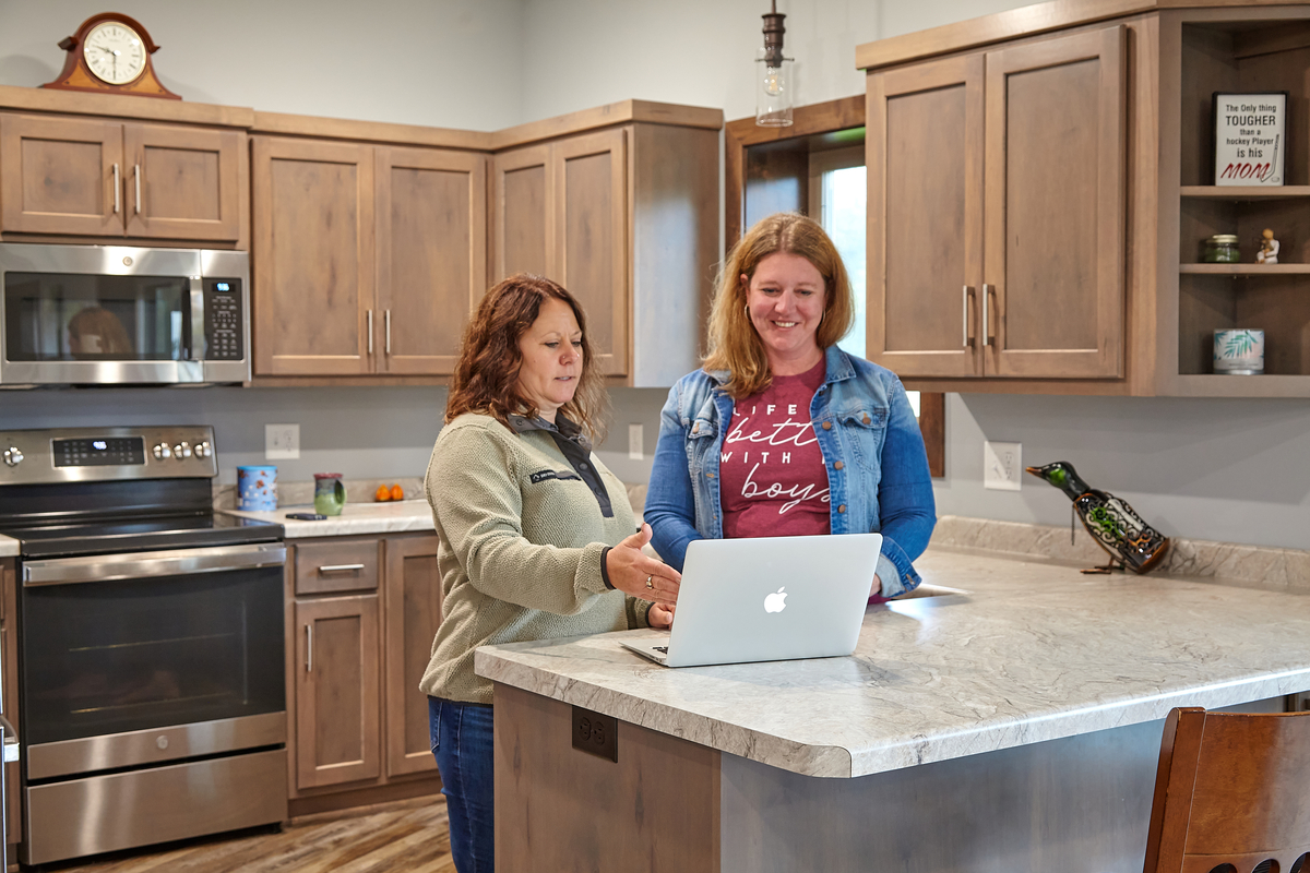 Two women standing at a kitchen island reviewing home project details together on a laptop.