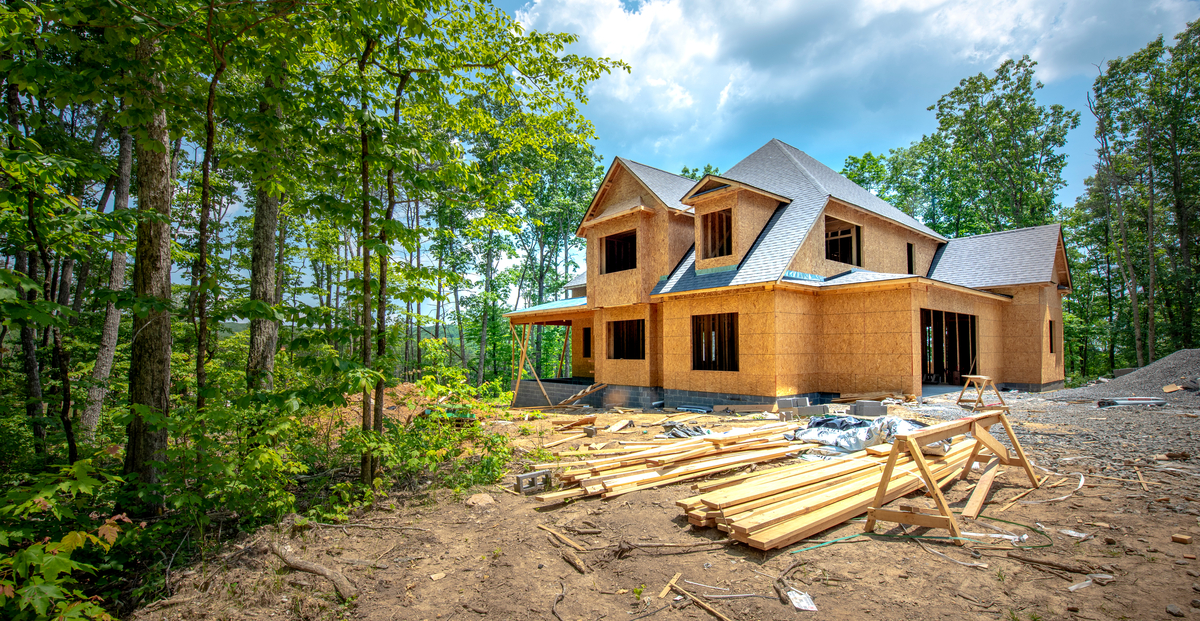 A large two-story house under construction with OSB sheathing stands in a wooded clearing under a bright, cloudy sky.