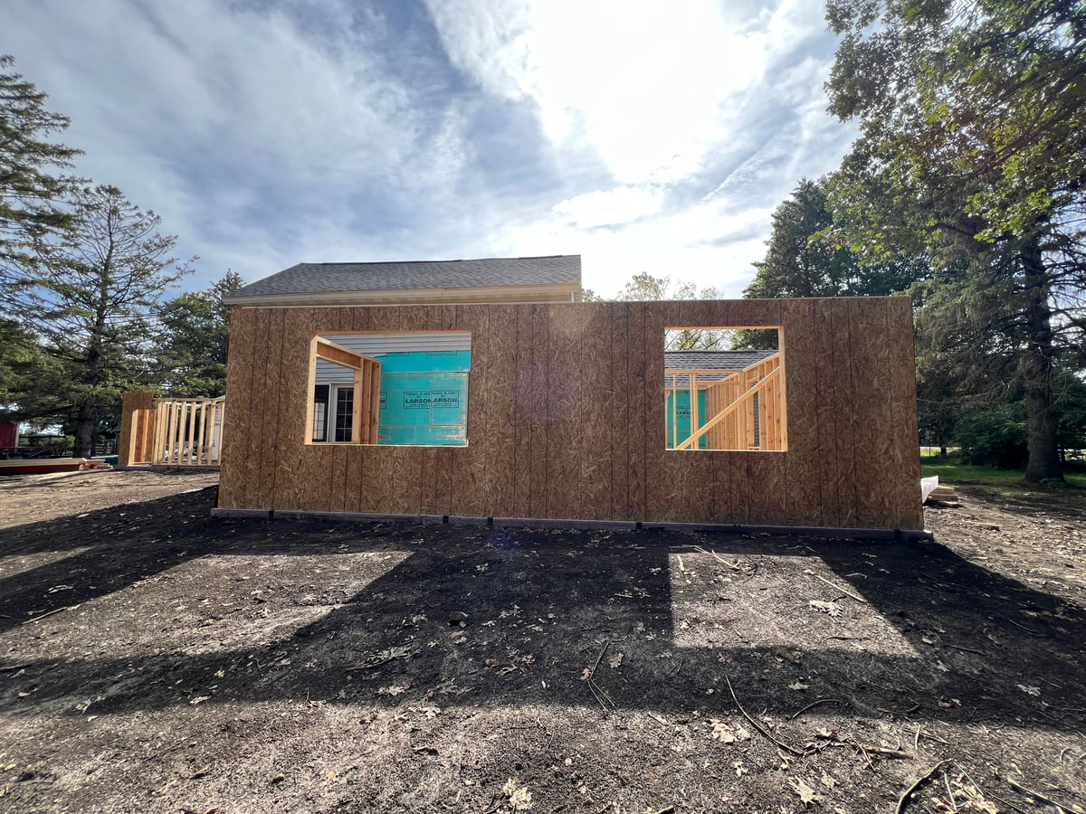 Exterior view of a home addition under construction with framed walls, window openings, and sheathing installed on a sunny day.