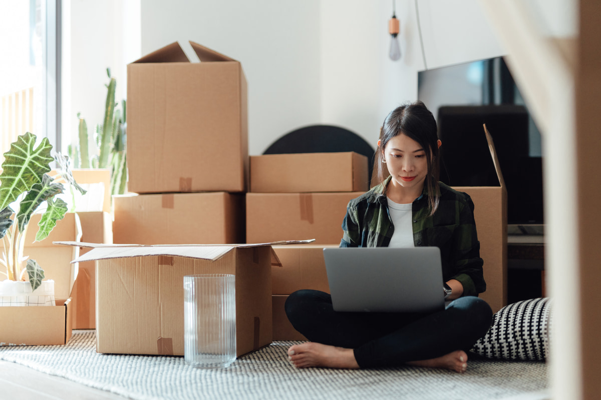 woman sitting on floor in front of boxes looking for new house on laptop