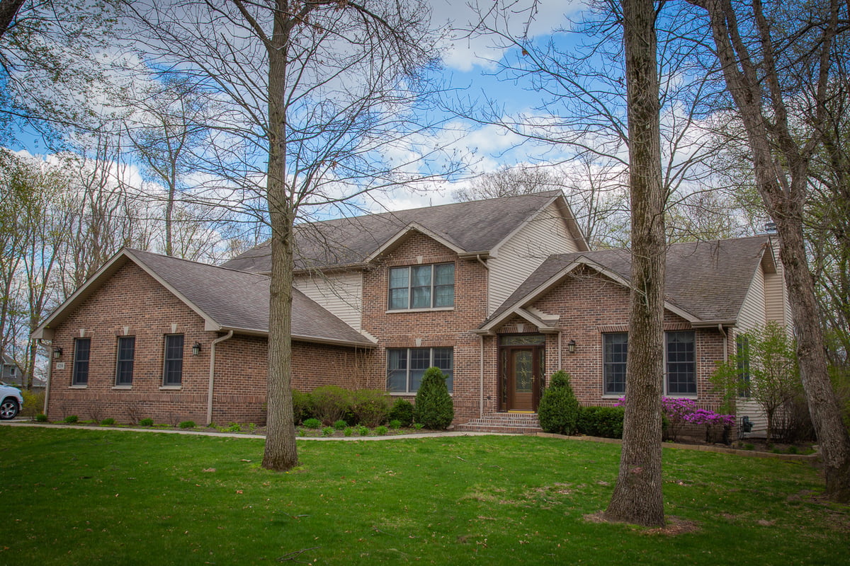 Brick two-story home with landscaped yard surrounded by tall trees in spring.
