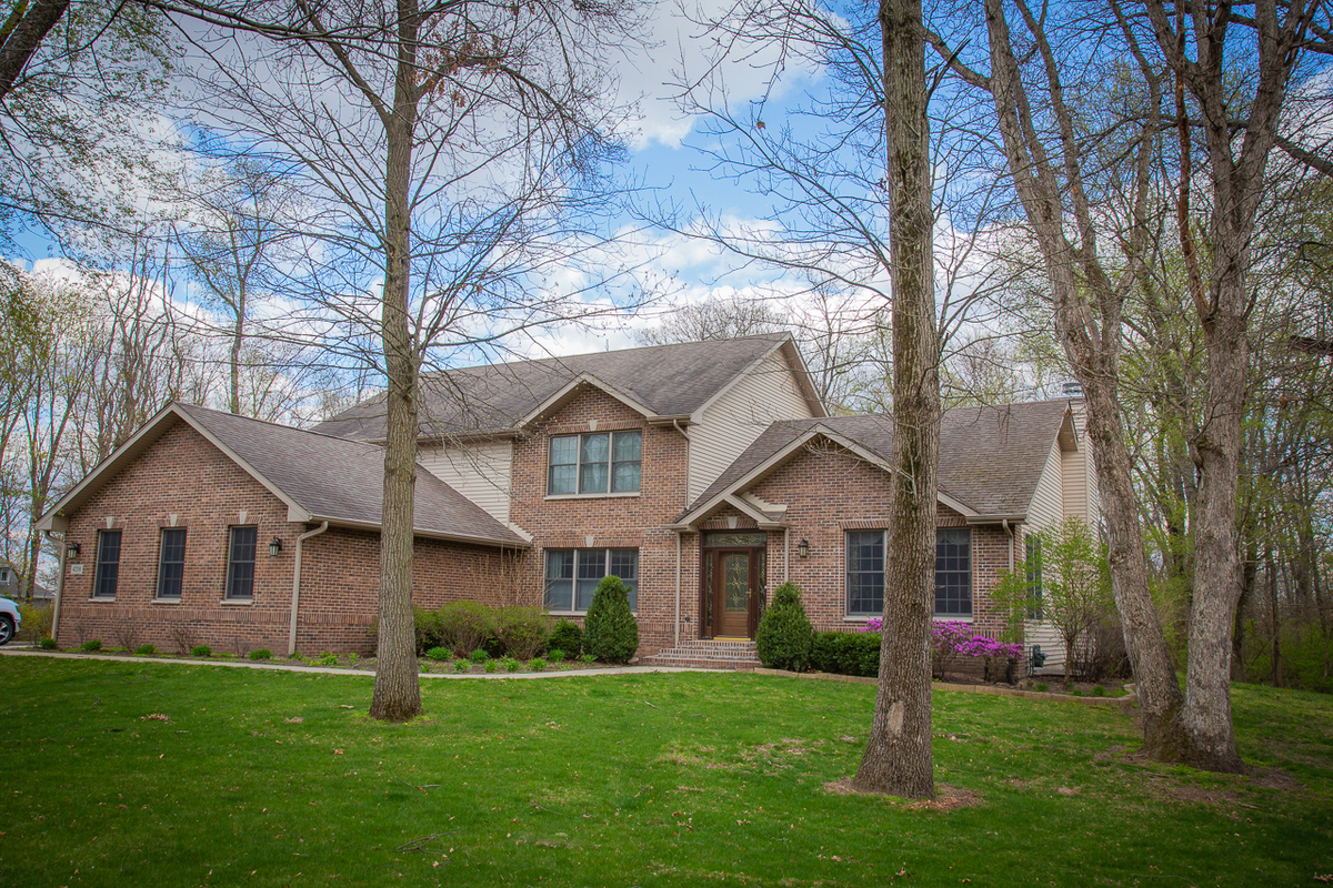 Brick two-story home with landscaped yard surrounded by tall trees in spring.
